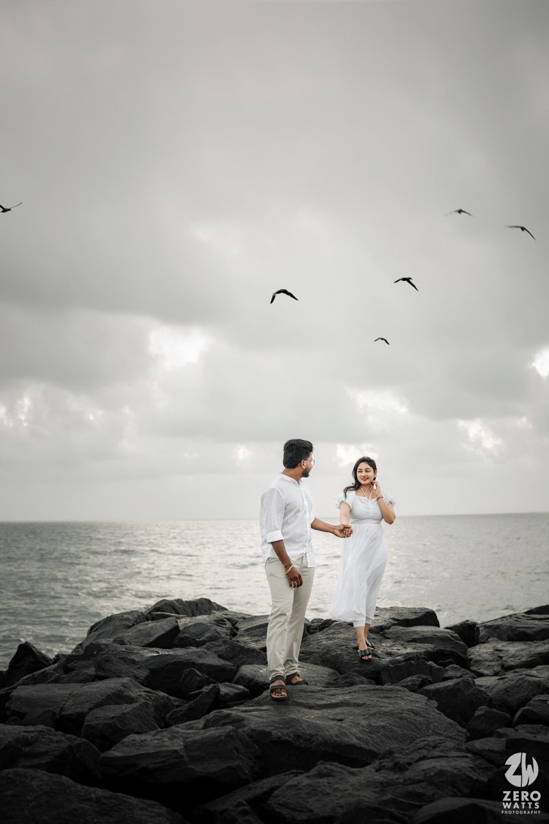 A couple stands together on dark rocky shoreline with seabirds flying overhead against moody gray sky.