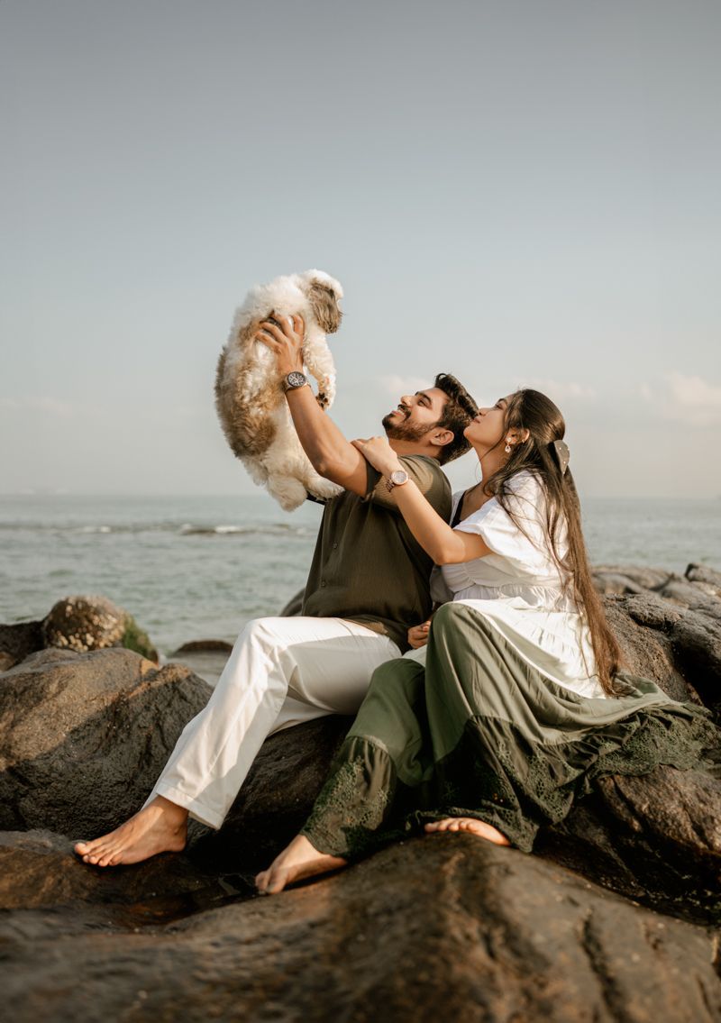 A couple sitting on coastal rocks lifting their small dog against a misty ocean background at sunset.