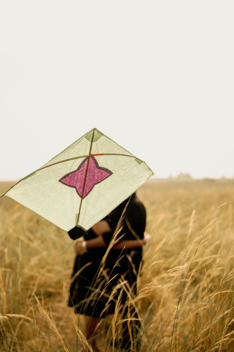 A silhouetted figure flies a simple white and pink kite in a golden wheat field at sunset.