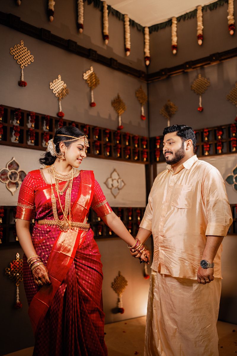 Couple in traditional South Indian wedding attire share a joyful moment under decorative hanging ornaments.