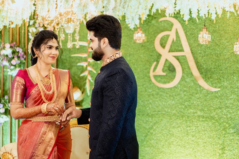 A couple in traditional Indian wedding attire standing in front of a decorative green backdrop with gold lettering.