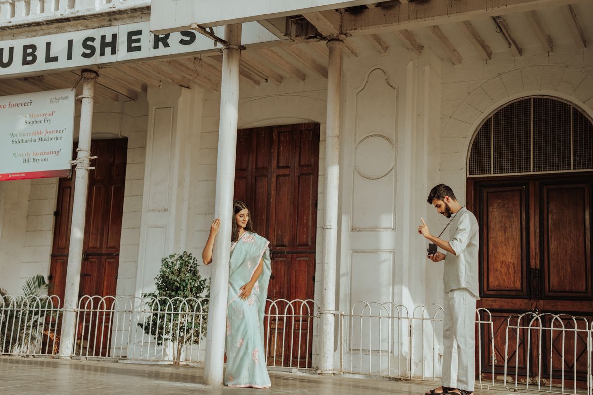 A couple in elegant attire stands apart on a vintage white colonial building porch with ornate railings and wooden doors.
