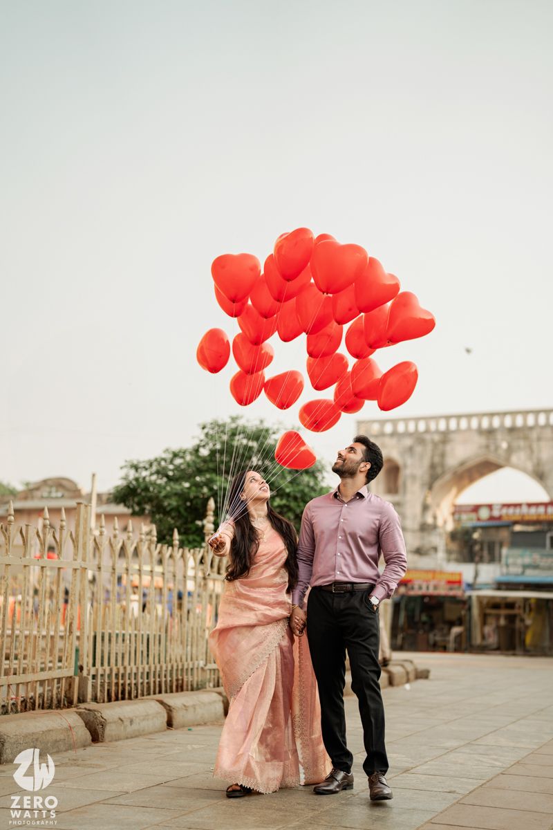 A couple holds red heart-shaped balloons against an overcast sky near historic architecture.