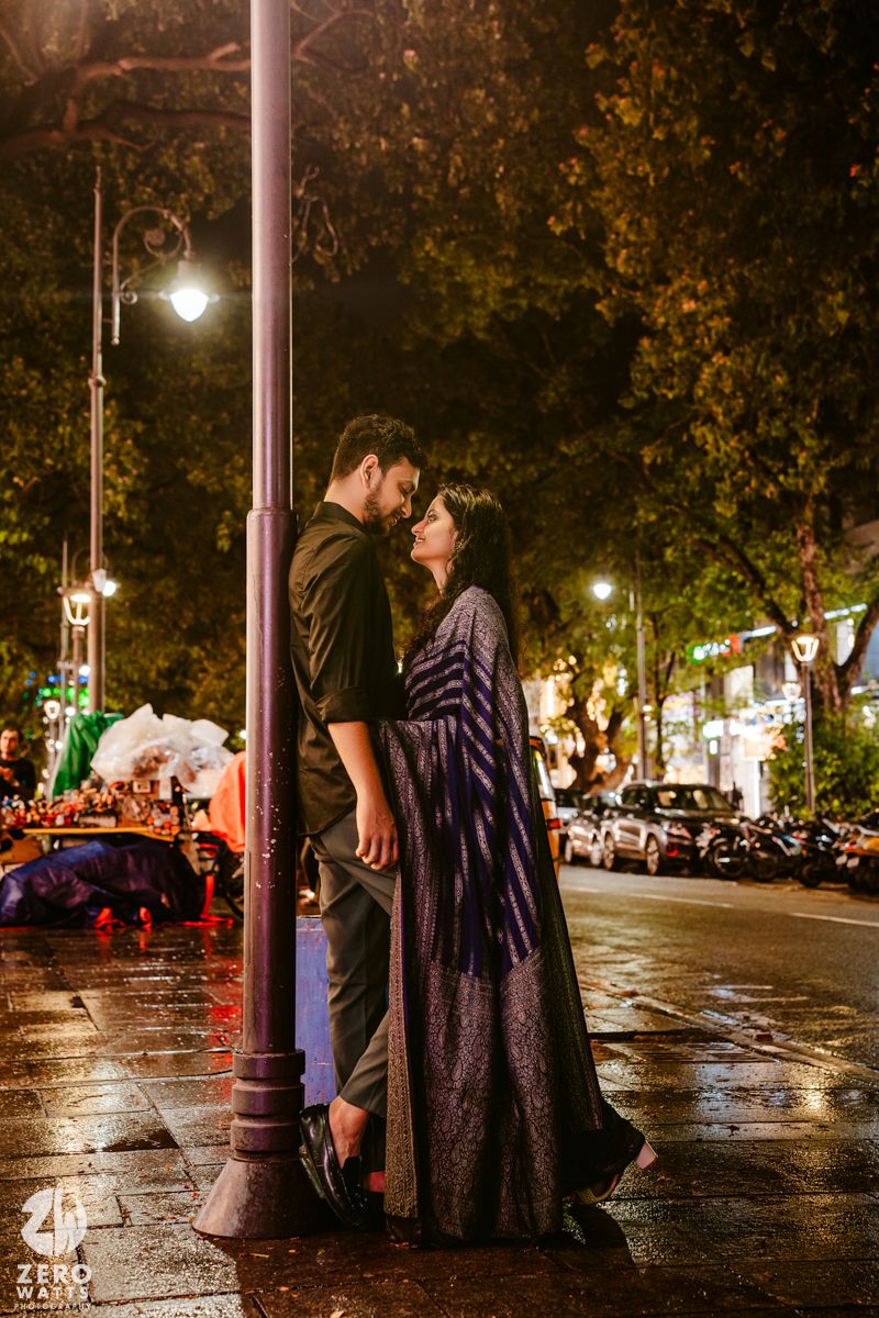 A romantic nighttime scene under streetlamp with wet pavement and city lights.