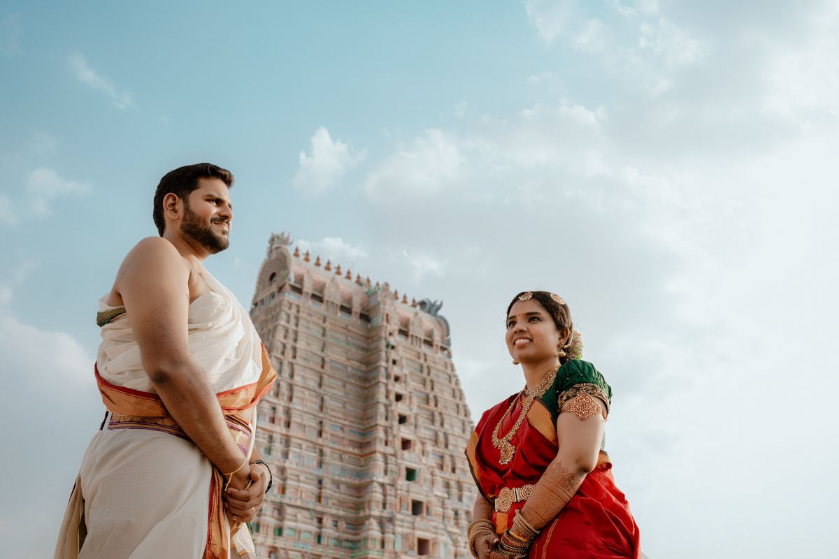 Two people in traditional South Indian attire stand in front of a temple gopuram against a cloudy sky.