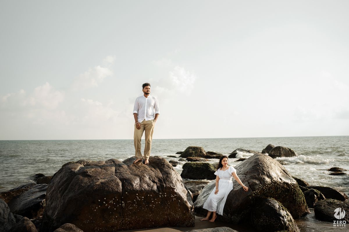 A couple poses on coastal rocks with ocean waves and cloudy sky in the background.