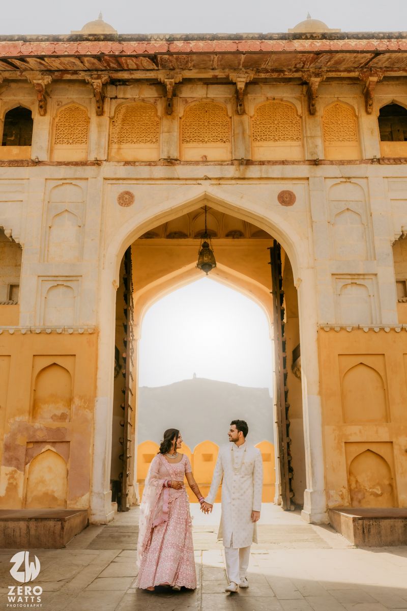 A couple in traditional Indian attire holding hands while walking through an ornate archway at sunset.