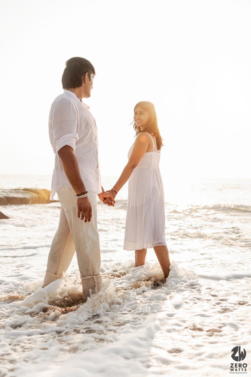 Two people in white outfits standing together in ocean waves during golden hour.