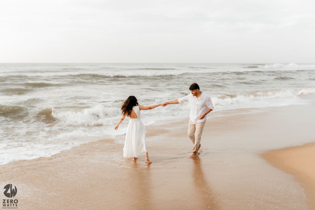 Two people in white outfits dance playfully along the shoreline as waves crash behind them.