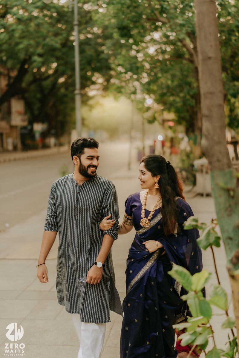 A couple wearing casual outfits walk together and share laughter on a tree-lined path on a sunny day.