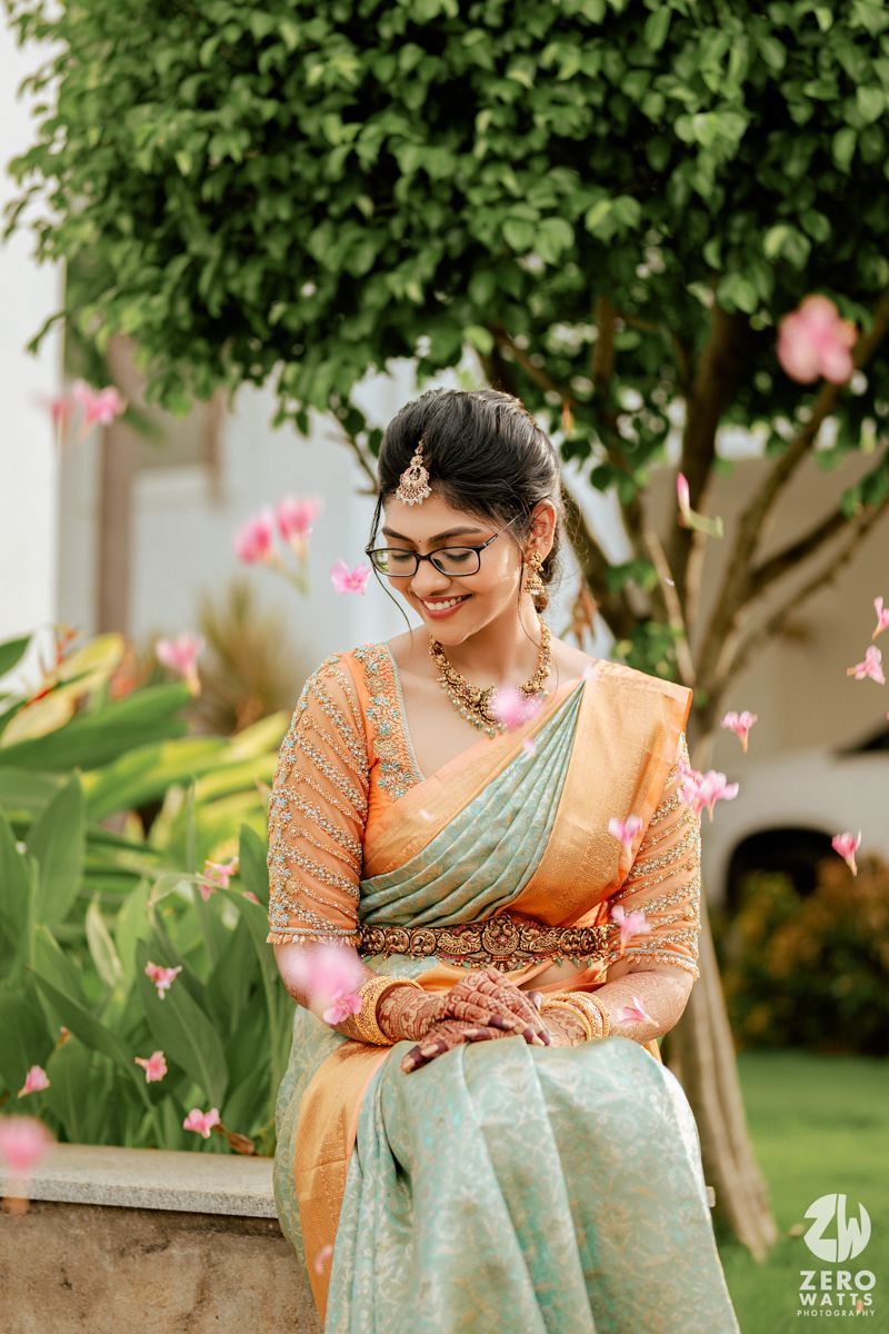 Person wearing orange and mint green traditional attire poses under flowering tree in garden setting.