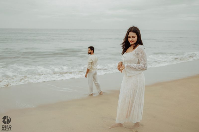 A couple in white outfits stands on a misty beach with ocean waves in the background.