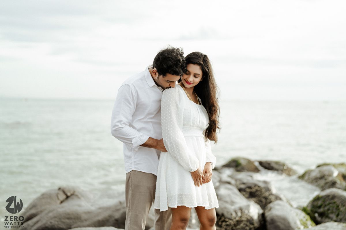A romantic couple dressed in white embraces on rocky seashore with ocean and cloudy sky in the background.