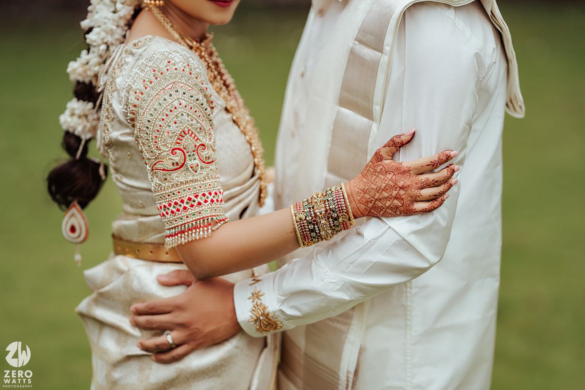 Close up detail of bride's hennaed hands and gold bangles on groom's white traditional outfit.