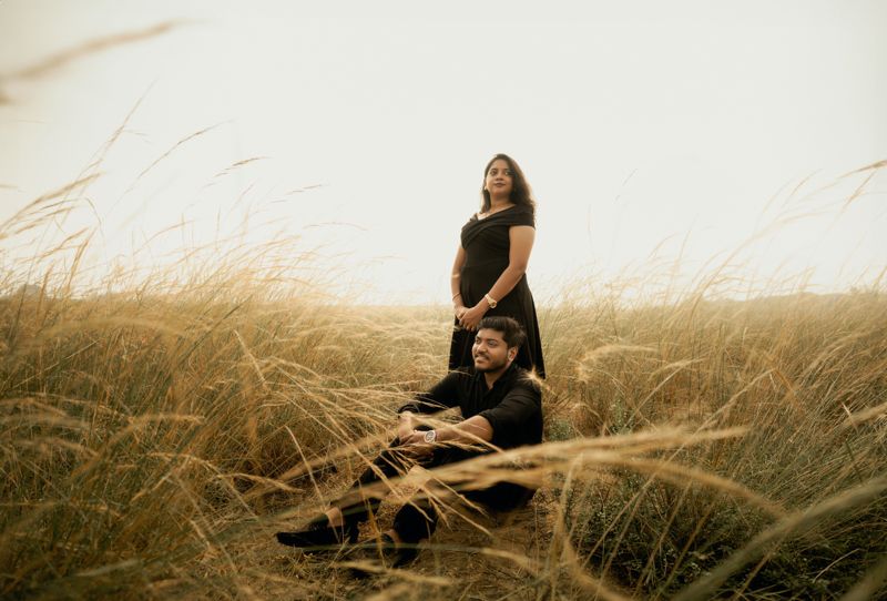 A couple sitting and standing in tall grass during golden hour in a dreamy, ethereal setting.