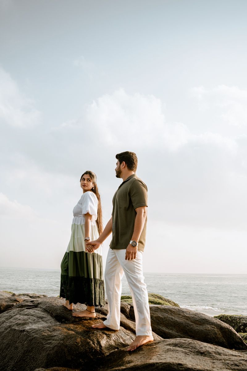A romantic couple standing together on coastal rocks overlooking the ocean at sunset.