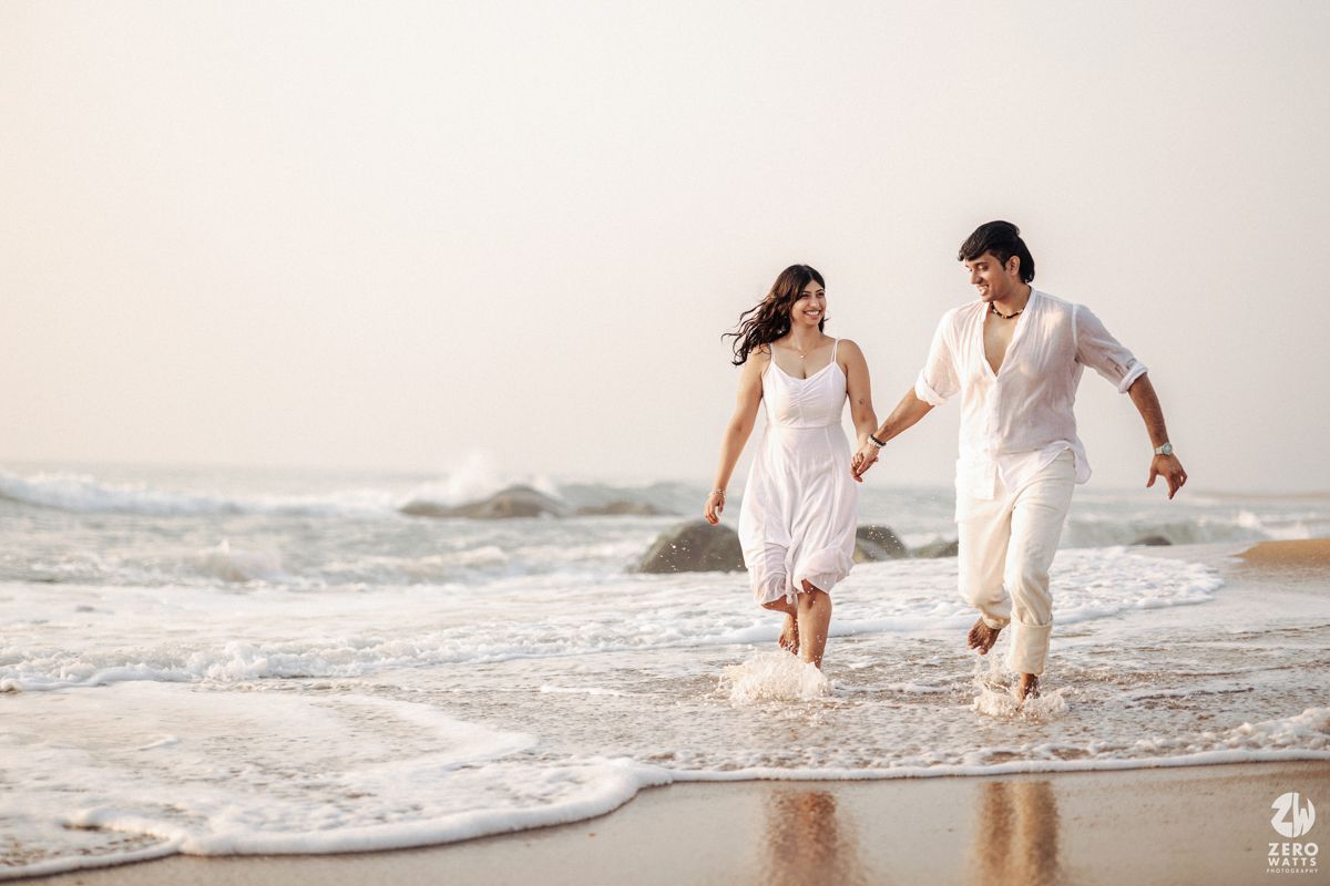A couple wearing white clothes playfully running through shallow ocean waves on a beach at dusk.