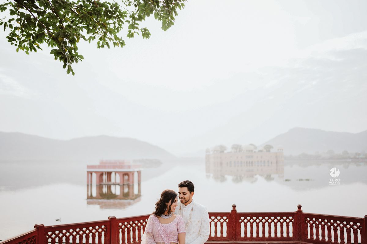 A couple in light-colored outfits stands on a red railing deck overlooking a misty lake with mountains in the background.