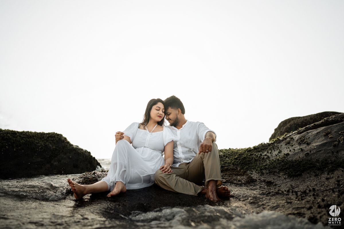 Couple in white outfits sit together on rocky coastal terrain.