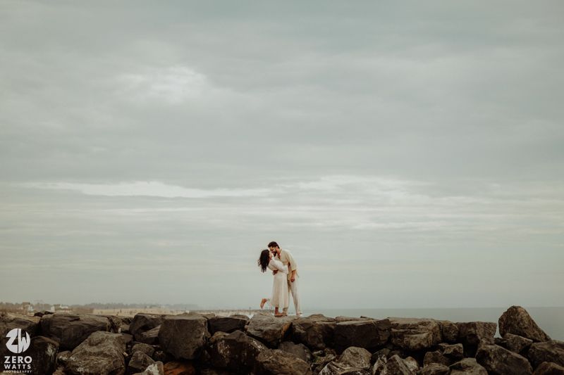 A couple embraces on rocky coastal outcrop against cloudy sky and ocean backdrop.