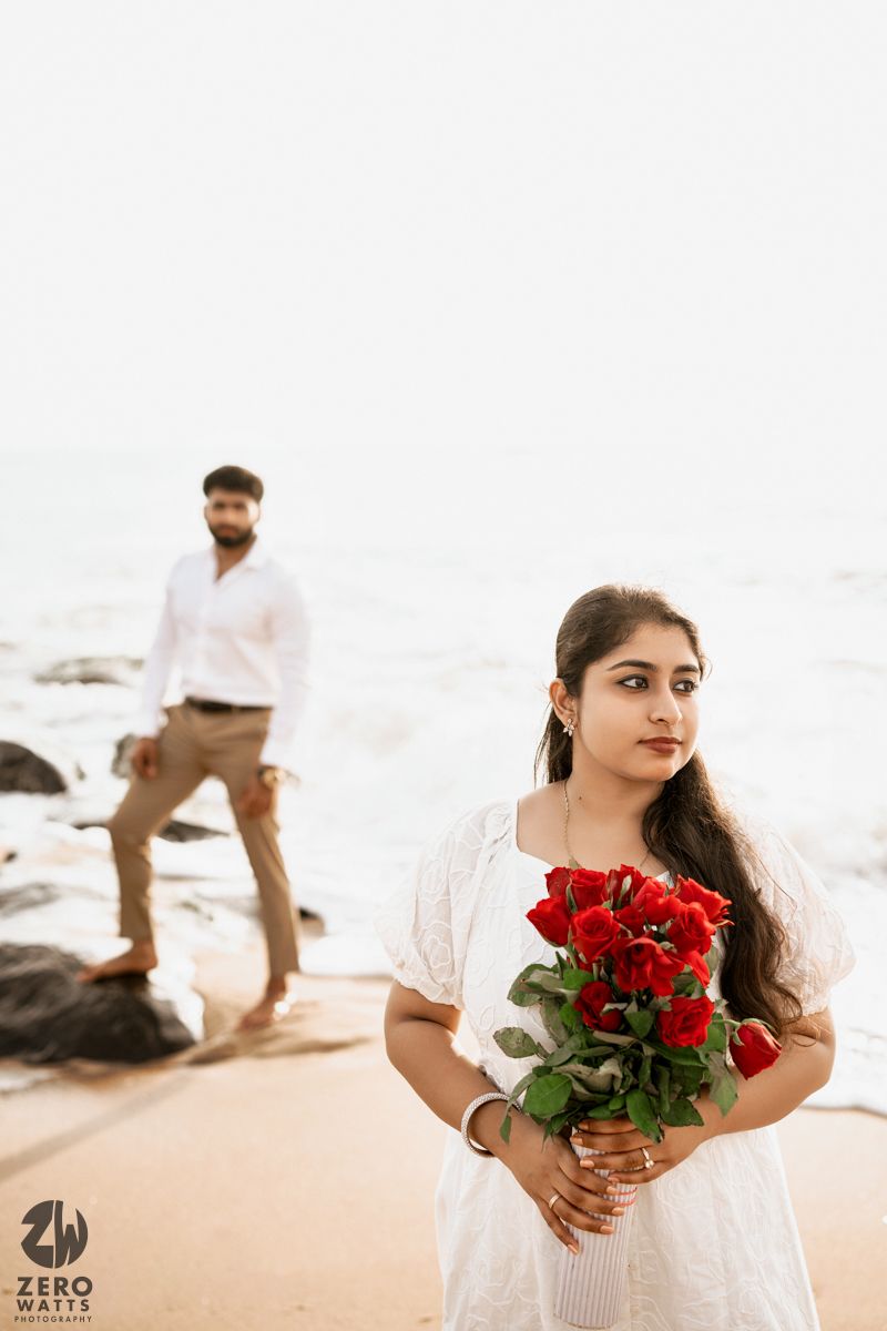 A woman in white dress holds red roses on beach while someone stands in background near rocks.