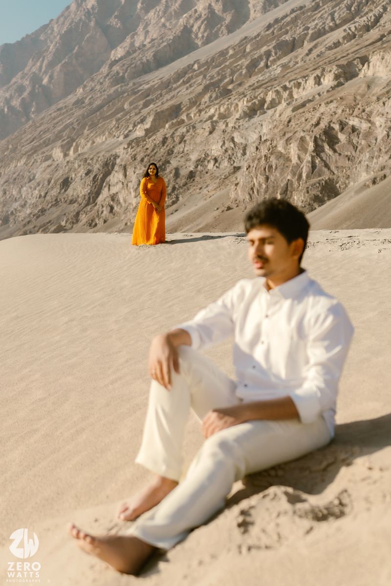 A figure reclines on a desert slope wearing a white outfit against a rocky mountainous backdrop at sunset.