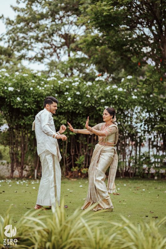 Couple holding hands and dancing in traditional beige and white attire outdoors.