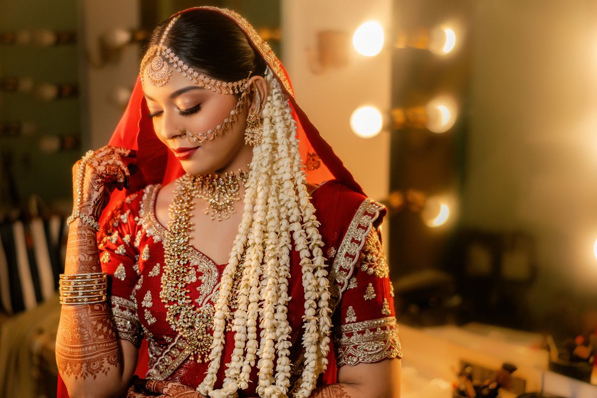 Traditional Indian bride wearing a red and gold lehenga with jasmine flowers and intricate jewelry in soft lighting.