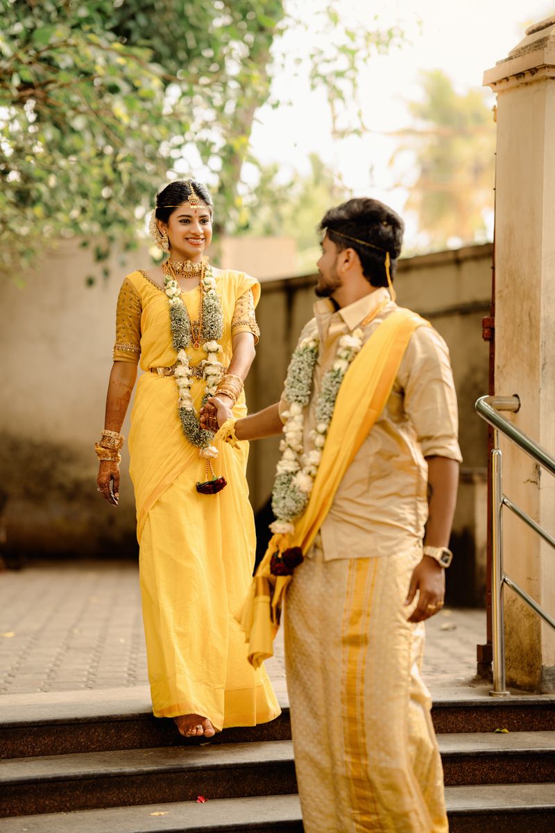 A couple in traditional yellow and cream attire stand on steps during a South Indian wedding ceremony.