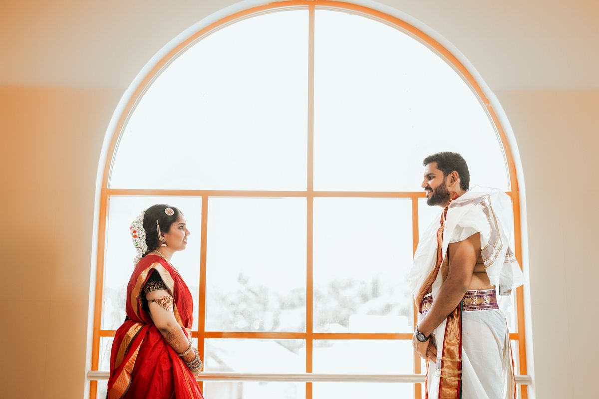 Two people in traditional Indian and modern attire stand facing each other in front of a large arched window.