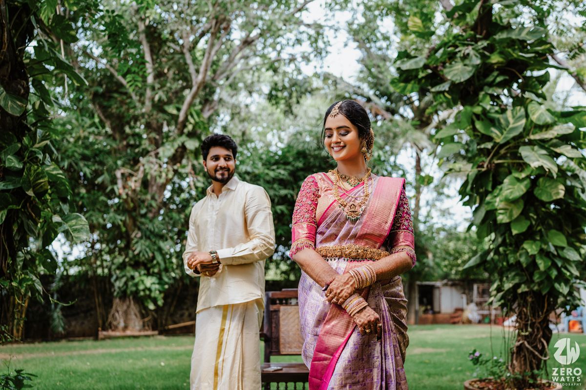 A couple in traditional South Indian wedding attire stands together in a garden setting with tall trees.