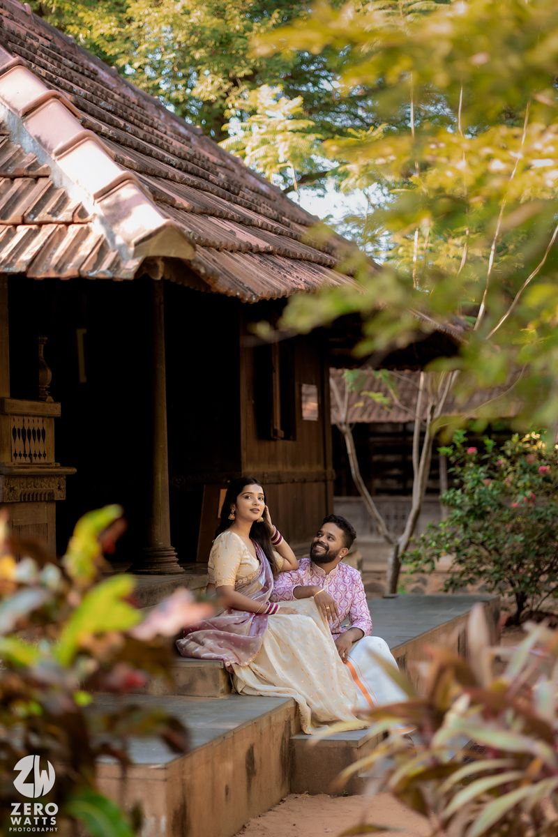 A couple sits together on the porch of a rustic wooden house with traditional tiled roof surrounded by autumn foliage.
