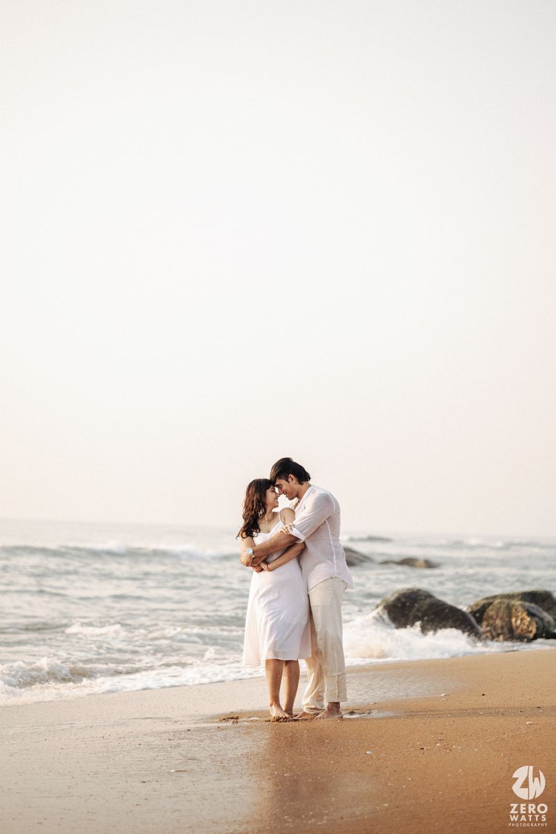 Romantic couple in white outfits shares intimate moment on sandy beach with crashing waves.