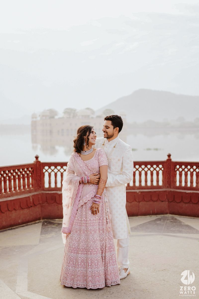 Couple in elegant pink and white traditional Indian wedding attire embracing on a terrace overlooking misty palace views.