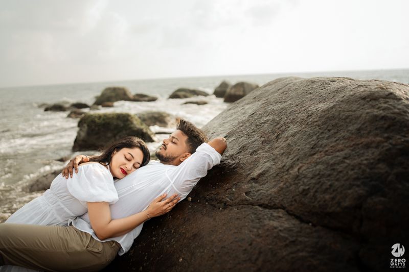 Couple relaxing on coastal rocks with waves and cloudy sky in the background.