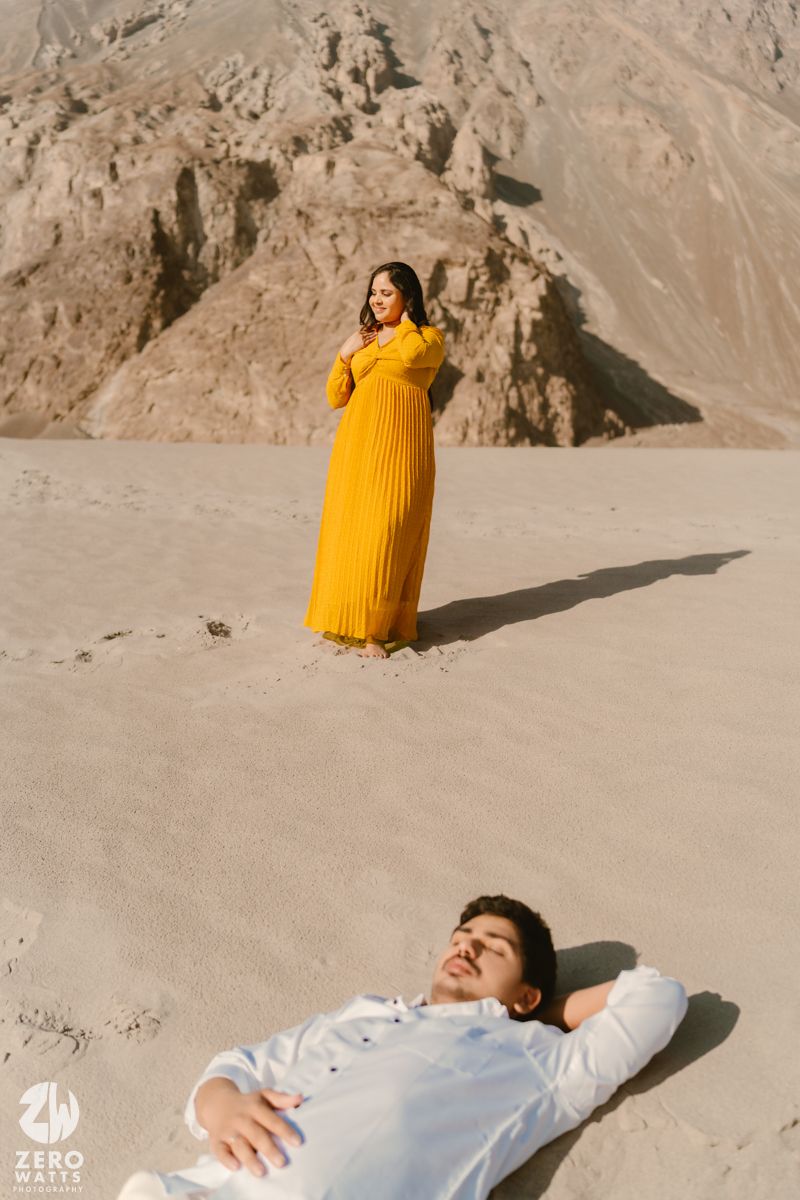 A wedding couple poses against a desert backdrop with the bride wearing a vibrant yellow gown and someone laying on the sand.
