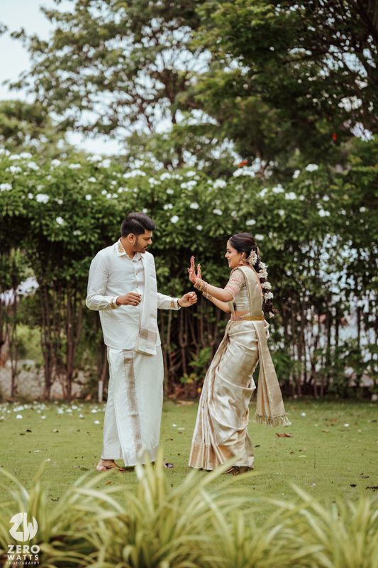 A couple in coordinating white and gold outfits dances playfully together on a grassy lawn with trees in the background.