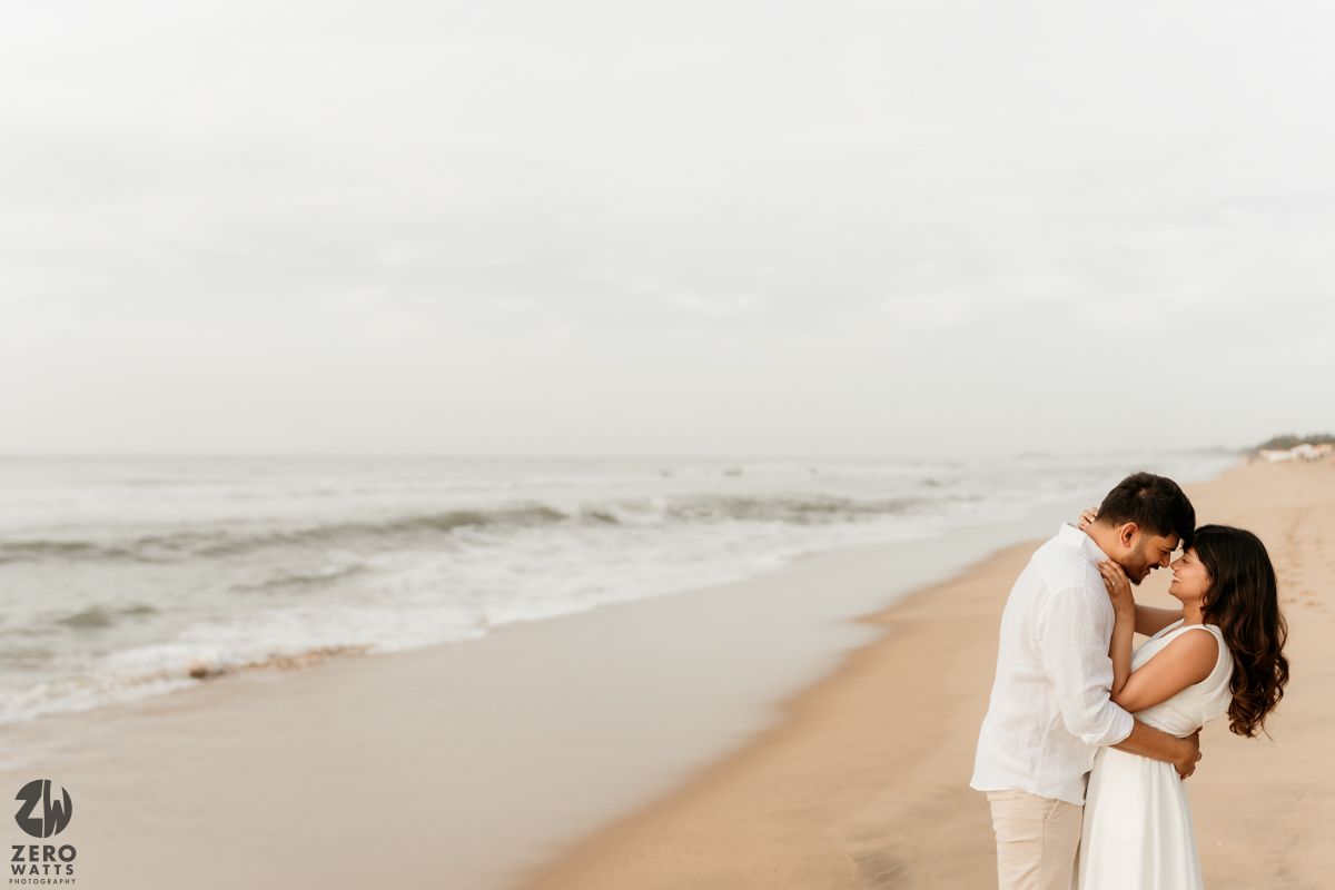 A romantic couple in white outfits embraces on a misty beach at sunset with waves crashing in the background.