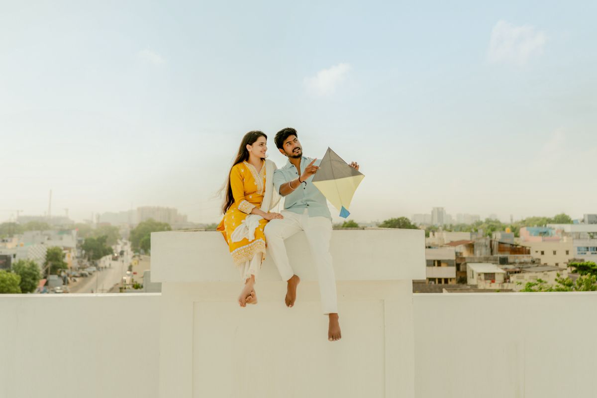 Two people wearing yellow and white sit reading a newspaper together on a white rooftop wall overlooking a city landscape.