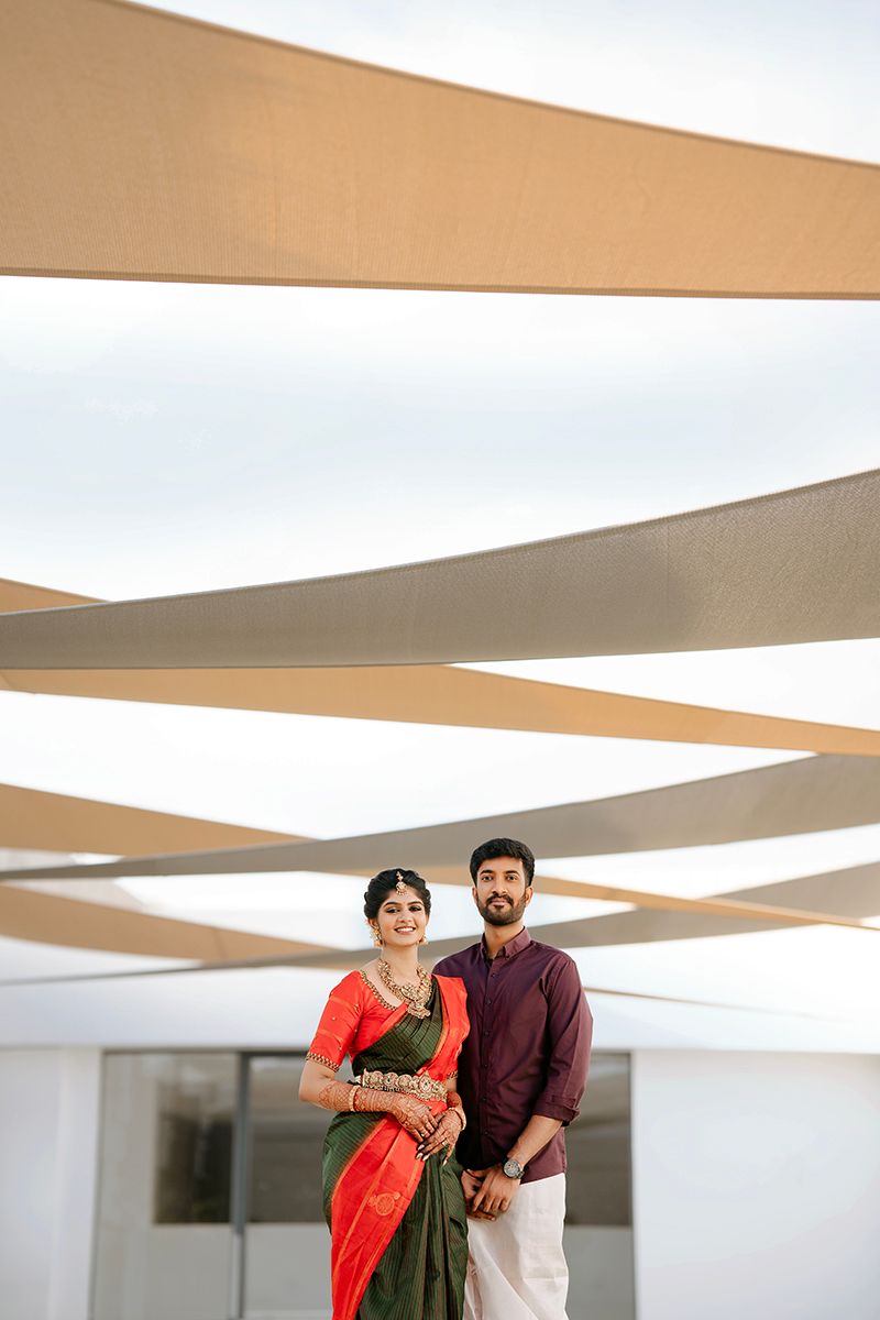 Couple stands together under modern architectural canopy with crossed beams while wearing traditional Indian attire.