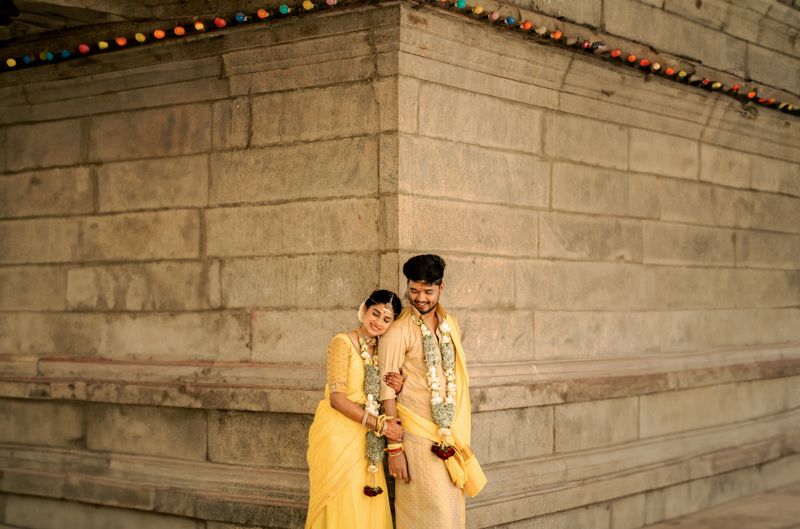 Two performers in yellow traditional outfits standing against a stone wall decorated with colorful lights.
