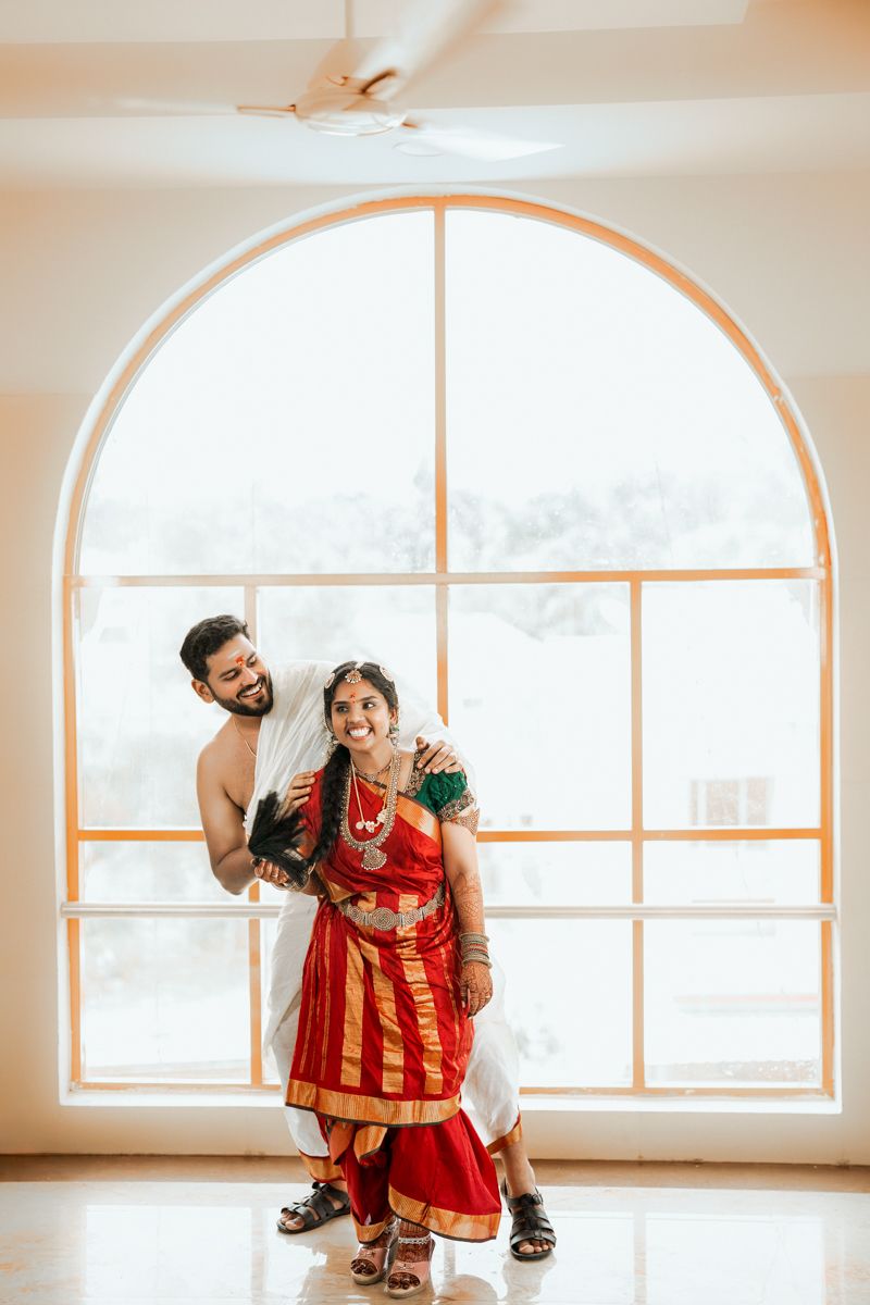 A couple dressed in traditional South Asian wedding attire shares a joyful moment in front of an arched window.