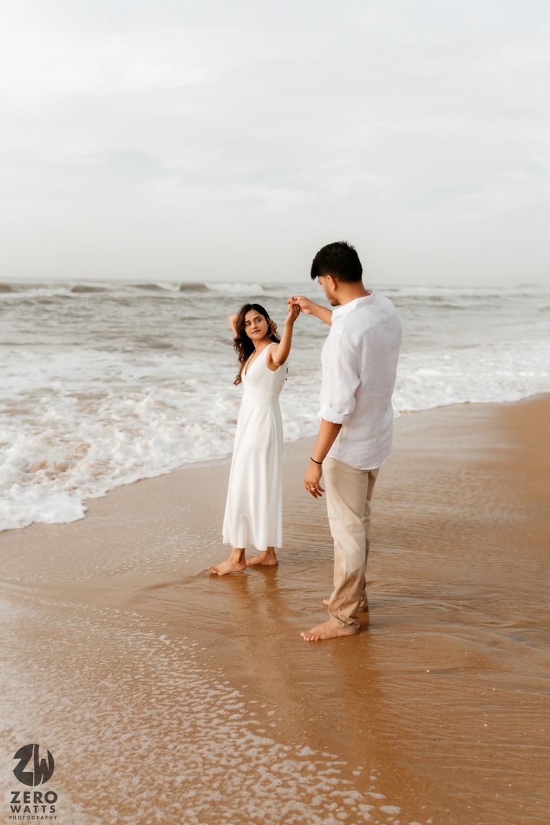 Romantic couple holds hands while walking along the shoreline during a beach photoshoot.