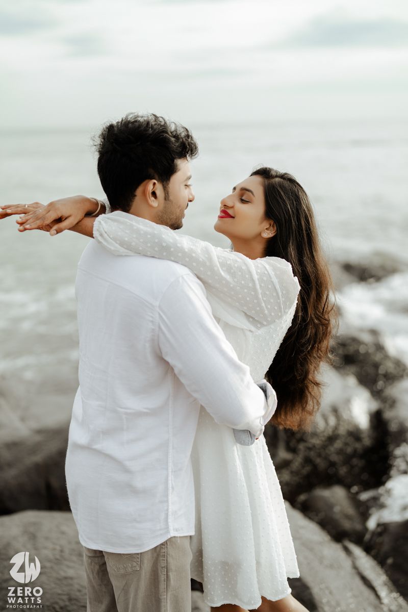 A romantic couple in white outfits embraces on coastal rocks by the ocean during an intimate photoshoot.
