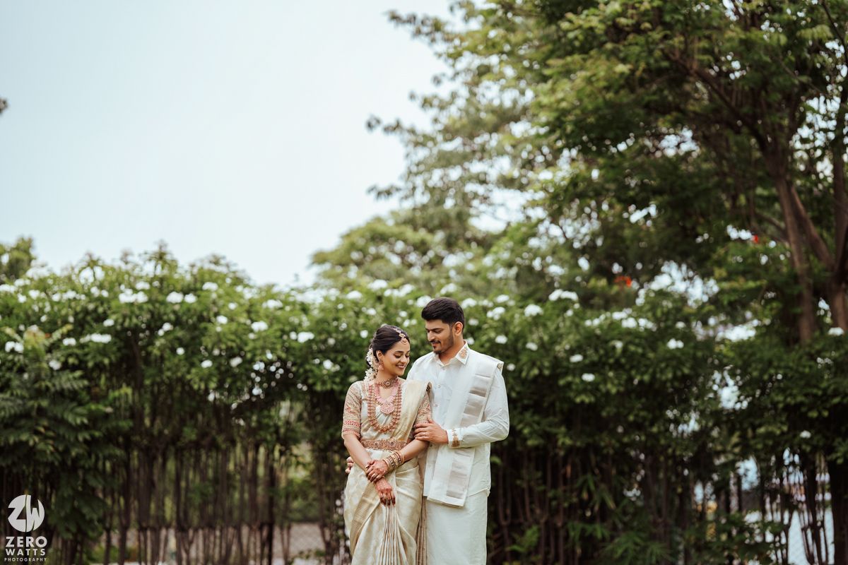 A couple in elegant cream-colored traditional Indian attire pose together in a lush garden setting.