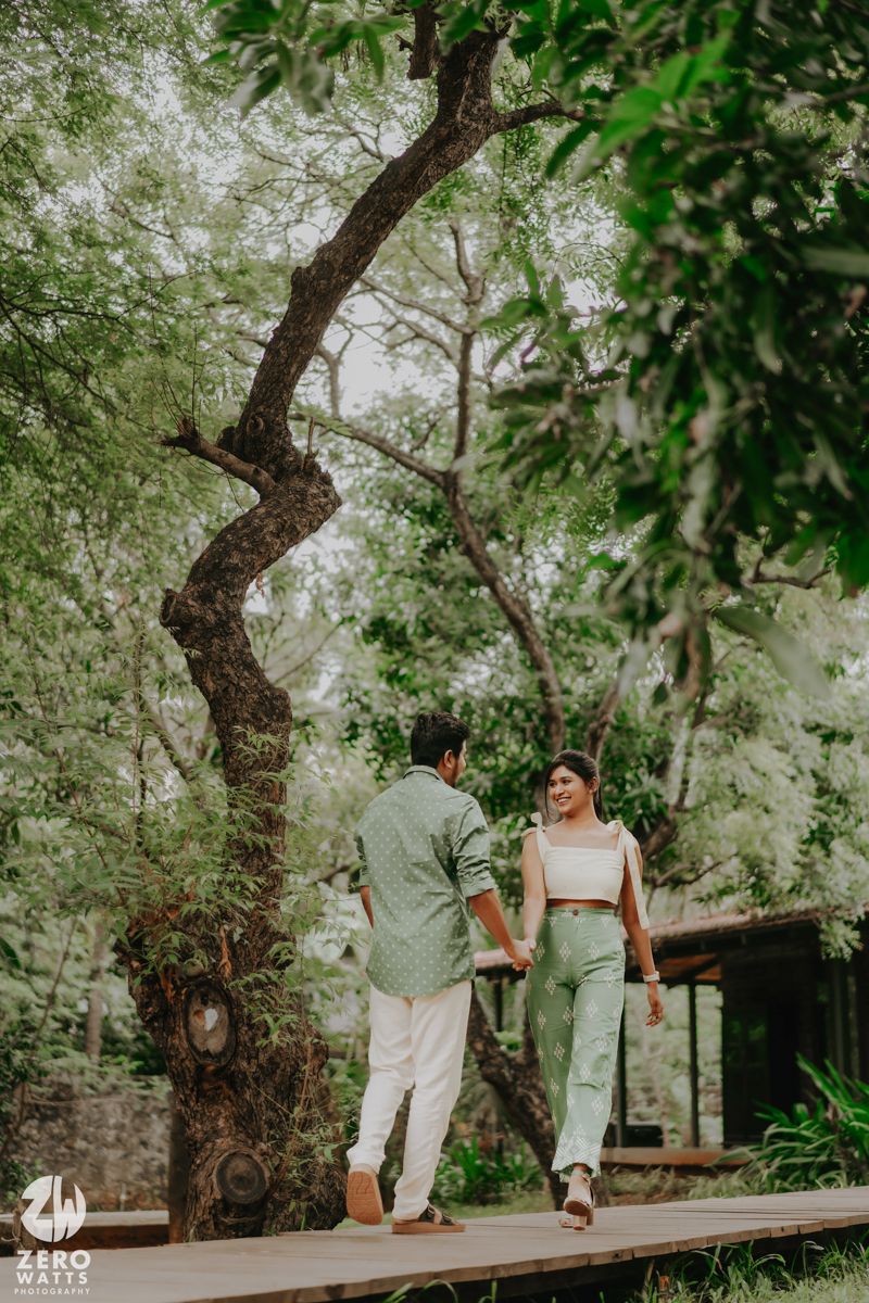 A couple walks hand in hand on a wooden pathway surrounded by lush green trees and twisted branches in a dreamy setting.