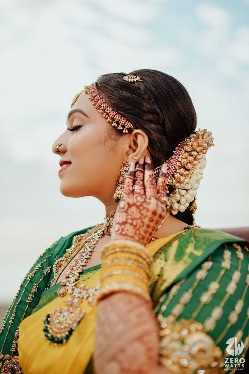 A traditional Indian bride in yellow and green attire adorned with jewelry and mehndi poses against a soft blue sky.