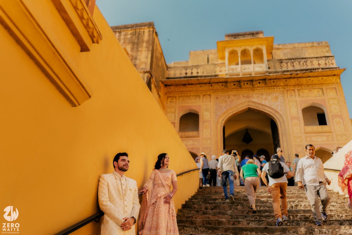 A couple stands against vibrant yellow walls at an ornate palace entrance while tourists descend stone steps in Jaipur.
