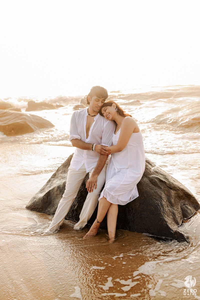 Couple in white clothing cuddles on large rock at beach during golden sunset.