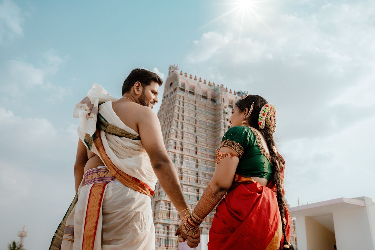 A couple in traditional Indian attire stands in front of a temple tower against a blue sky.